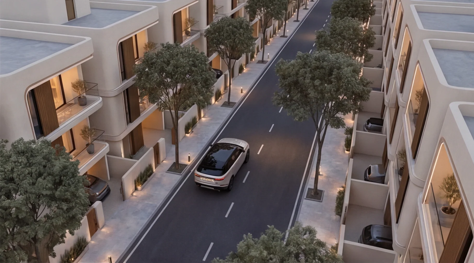 A white car driving down a tree-lined internal street at dusk, seen from above, with villas and lit boundary walls on both sides
